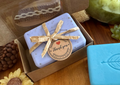 A gift box of artisanal soaps displayed on a wooden table. The box is open to reveal a lavender soap in a clear window and a lavender soap tied with a twine bow and a 'Thank you' tag.